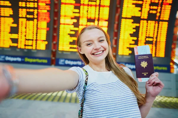 Lány nemzetközi repülőtéren, figyelembe véve a vicces selfie a passport és a beszállás át közelében repülési információs tábla — Stock Fotó