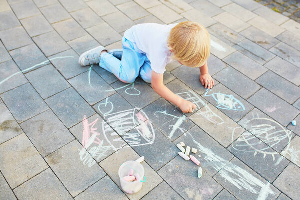 Adorable little boy drawing with colorful chalks on asphalt. Summer activity and creative games for small kids