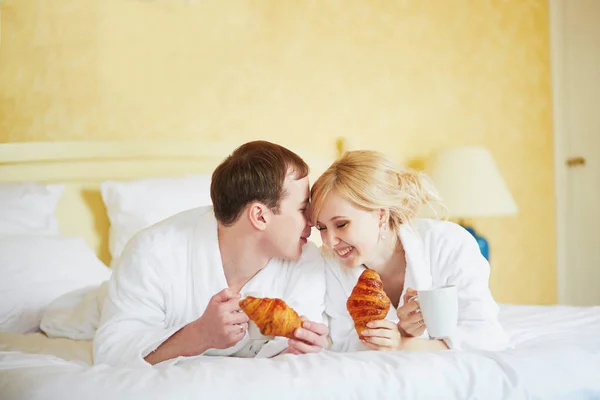 Jovem Casal Feliz Roupões Banho Brancos Comendo Croissants Juntos Cama — Fotografia de Stock