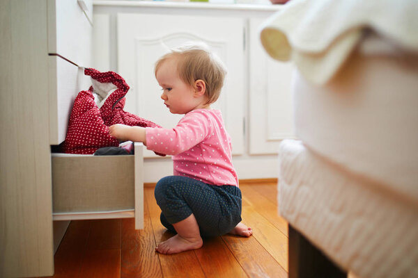 Adorable toddler girl at home, opening the drawer of dresser and taking out clothes. Curious little kid exploring her flat or house. Home child proofing concept