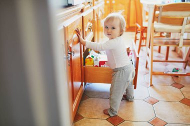 Adorable toddler girl at home, opening the drawer in the kitchen and selecting food. Curious little kid exploring her flat or house. Home child proofing concept clipart
