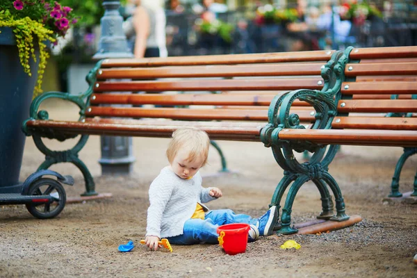 フィンランドのヘルシンキの通りのベンチの近くの地面に座っている小さな赤ん坊の女の子 幼児砂型 バケツとシャベルで遊んで 小さな石を集めてください 子供のための屋外創造活動 — ストック写真