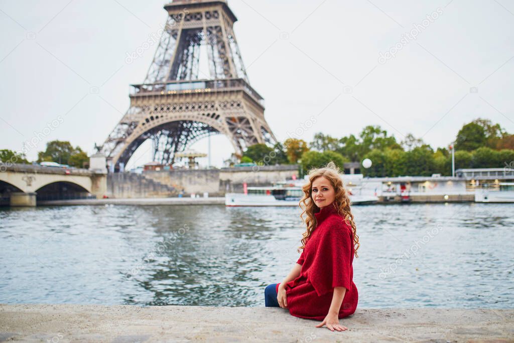 Young woman with long blond curly hair in Paris, France. Beautiful tourist in red coat near the Eiffel tower, on the Seine embankment