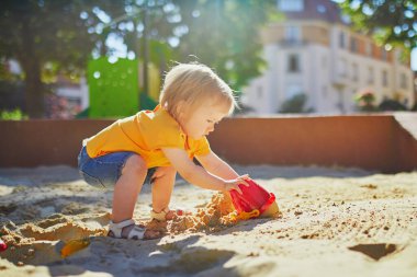 Sevimli küçük kız kum havuzunda oyun parkında eğleniyor. Bebek kum kalıplarıyla oynuyor ve çamur yapıyor. Çocuklar için açık hava yaratıcı aktiviteler