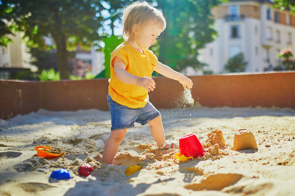 Очаровательная маленькая девочка развлекается на детской площадке в песочнице. Тоддлер играет с песчаными плесенями и делает грязи. Активный отдых на свежем воздухе для детей