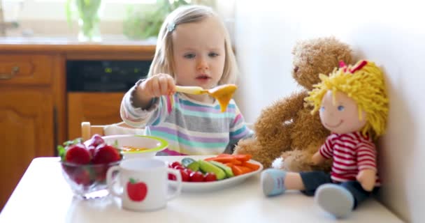 Adorable niña pequeña comiendo frutas y verduras frescas para el almuerzo. Muñeca de alimentación infantil y oso de peluche en la cocina. Deliciosa comida saludable para niños — Vídeos de Stock