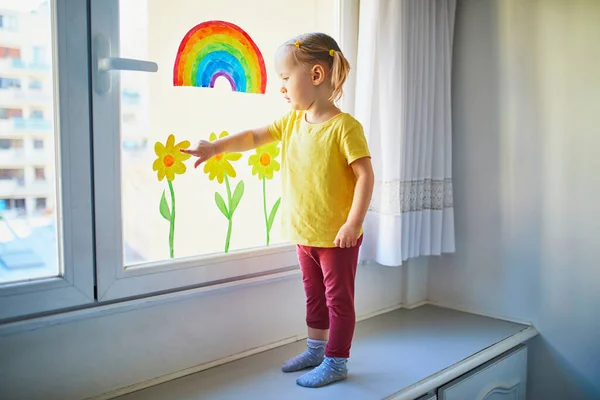 Adorable toddler girl attaching drawing of rainbow to window glass as sign of hope. Creative games for kids staying at home. Self isolation and quarantine concept