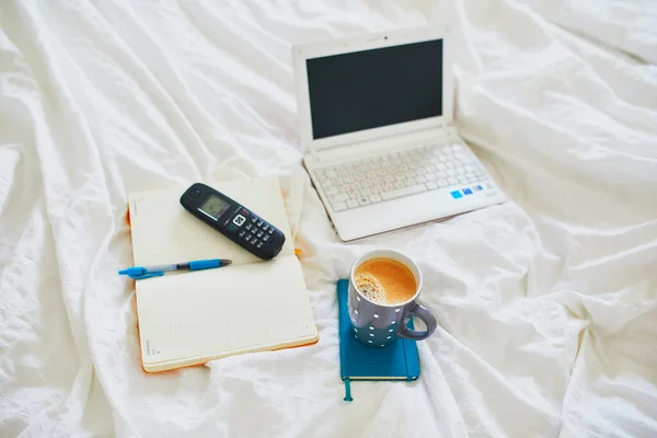 Laptop, cup of coffee, notebook and phone in bed on clean white linens. Freelance, distance learning or work from home concept