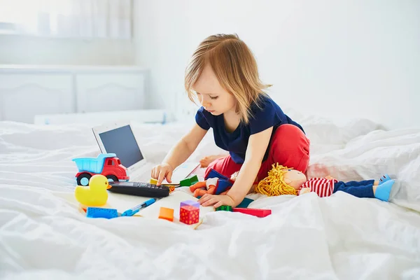 一个带着笔记本电脑 笔记本 电话和不同玩具躺在干净的白布床上的小女孩 自由学习 远距离学习或在家带孩子工作的概念 — 图库照片