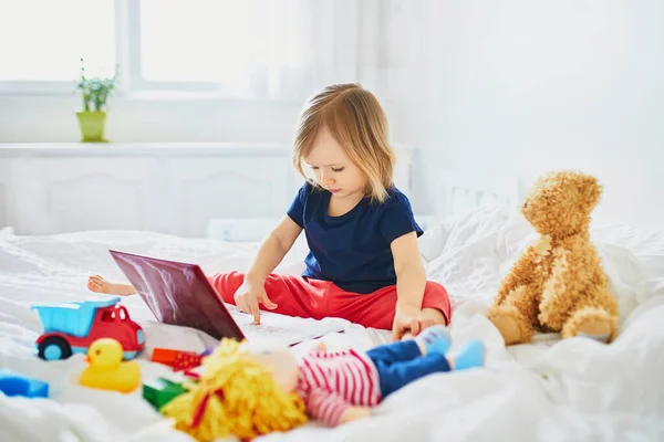 Toddler girl with laptop and toys in bed. Kid using gadget to communicate with friends or kindergartners. Education, distance learning or work from home with kids. Stay at home entertainment