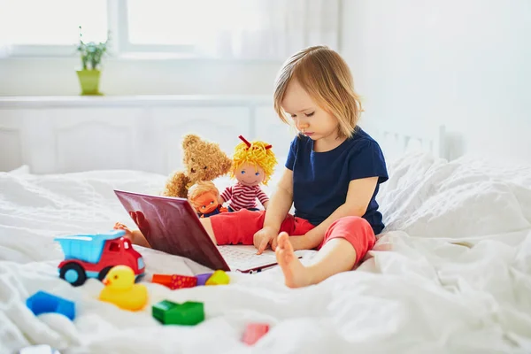 ベッドの中でラップトップとおもちゃと幼児の女の子 友達や幼稚園生とコミュニケーションするためにガジェットを使用して子供 子供と一緒に家から教育 距離学習や仕事 ホームエンターテイメントに滞在する — ストック写真