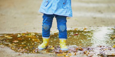 Sarı çizme giyen ve sonbahar günü su birikintisine atlayan bir çocuk. Küçük kız yağmurlu bir günde parkta su ve çamurla eğleniyor. Çocuklar için açık hava sonbahar aktiviteleri