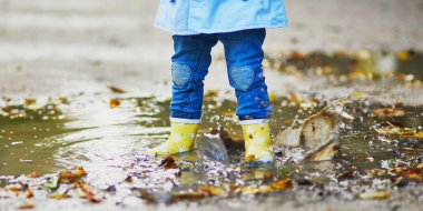 Child wearing yellow rain boots and jumping in puddle on a fall day. Toddler girl having fun with water and mud in park on a rainy day. Outdoor autumn activities for kids clipart