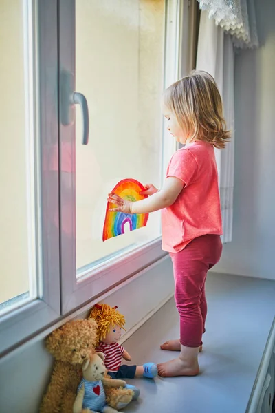 可爱的蹒跚学步的小女孩把彩虹画绑在窗玻璃上作为希望的象征 孩子们在关门大吉时呆在家里的创意游戏 自我隔离和验尸官检疫概念 — 图库照片