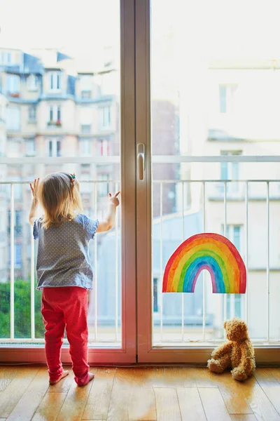 可爱的蹒跚学步的小女孩 带着彩虹的图画从窗户往里看 孩子们在关门大吉时呆在家里的创意游戏 自我隔离和验尸官检疫概念 — 图库照片