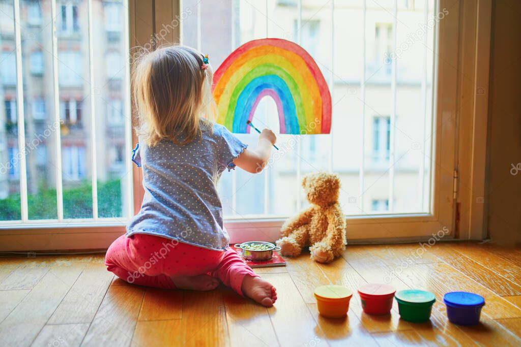 Adorable toddler girl painting rainbow on the window glass as sign of hope. Creative games for kids staying at home during lockdown. Self isolation and coronavirus quarantine concept