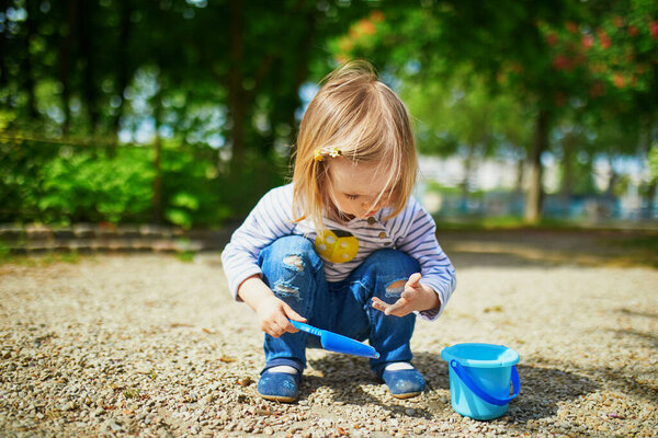 Очаровательная девочка играет с ведром и лопатой, делает грязь и собирает маленькие камни. Активный отдых на свежем воздухе для детей