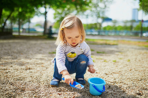 Очаровательная девочка играет с ведром и лопатой, делает грязь и собирает маленькие камни. Активный отдых на свежем воздухе для детей