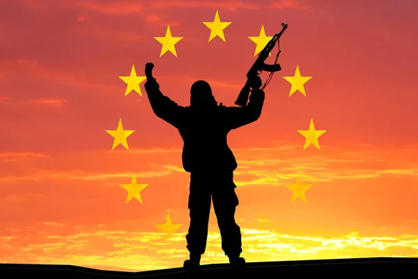 Silhueta de soldado militar, tiro, segurando arma, céu colorido, conceito de um terrorista. Terroristas silhuetas com espingarda, bandeira nacional em segundo plano - União Europeia - UE — Fotografia de Stock