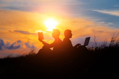 Dizüstü bilgisayarı olan bir adamın silueti ve günbatımı veya gündoğumu arka planı üzerine bir kitap.
