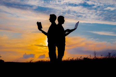 Dizüstü bilgisayarı ve günbatımında ya da gündoğumunda bir kitabı olan bir adamın silueti. 