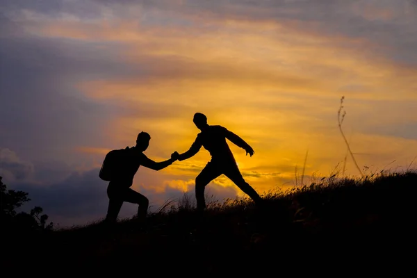 Caminhadas de casal de trabalho em equipe ajudam uns aos outros a confiar na silhueta de assistência nas montanhas, pôr do sol. Trabalho em equipe de dois homens caminhantes ajudando uns aos outros no topo da equipe de escalada, bela paisagem do pôr do sol . — Fotografia de Stock