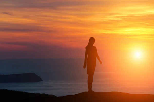 Silueta de una niña de pie en una colina mirando directamente al fondo de una hermosa puesta de sol y el mar —  Fotos de Stock