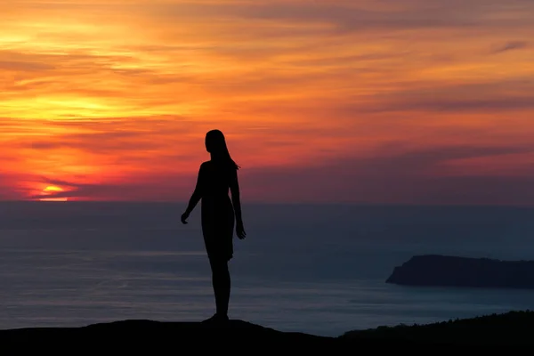 Silueta de una niña de pie en una colina mirando directamente al fondo de una hermosa puesta de sol y el mar —  Fotos de Stock