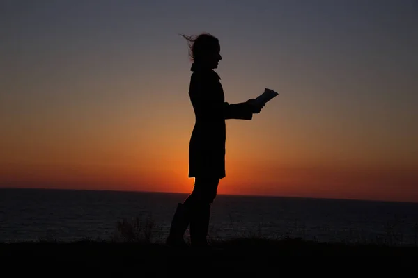 Silueta de una niña leyendo un interesante libro sobre el fondo de una hermosa puesta de sol — Foto de Stock