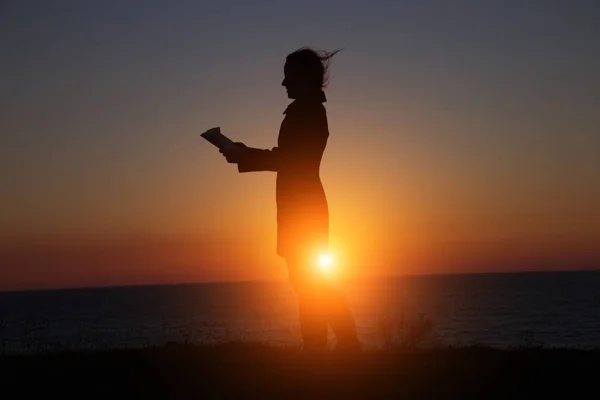 Silueta de una niña leyendo un interesante libro sobre el fondo de una hermosa puesta de sol —  Fotos de Stock