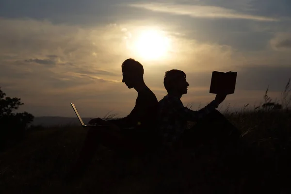 Dos amigos sentados en la colina de la lectura de un libro y sentados en un ordenador portátil en el fondo de una hermosa puesta de sol — Foto de Stock