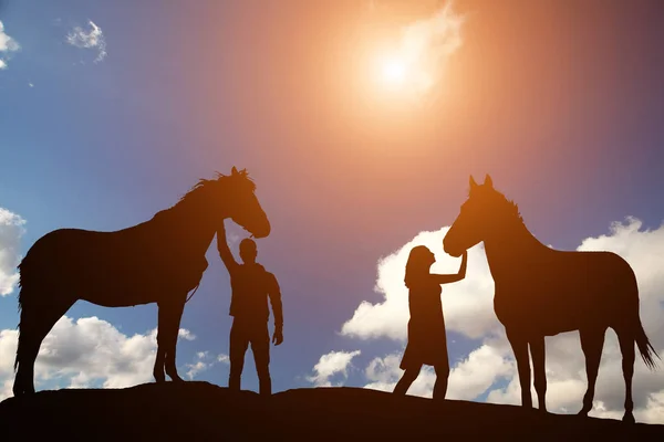 Silueta chico y chica acarició dos caballos en un fondo de una hermosa puesta de sol —  Fotos de Stock