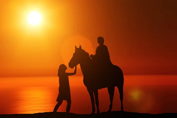 Silhouette of a man sitting on a horse standing next to a girl on the background of a beautiful sunset — Stock Photo, Image