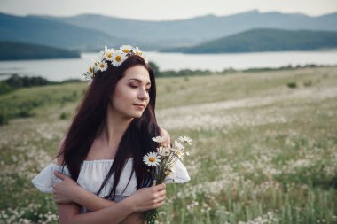 Beyaz elbiseli güzel esmer kadın ve çiçek çelengi papatya tarlasında baharın tadını çıkarıyor. Sağlık fikri ve konsept, güzel sağlıklı saç ve deri, alerji eksikliği.