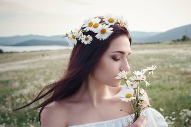 Beyaz elbiseli güzel esmer kadın ve çiçek çelengi papatya tarlasında baharın tadını çıkarıyor. Sağlık fikri ve konsept, güzel sağlıklı saç ve deri, alerji eksikliği.