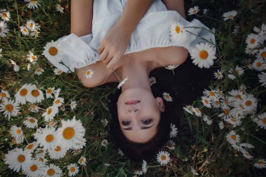 Beyaz elbiseli güzel esmer kadın papatya tarlasında baharın tadını çıkarıyor. Güzel bir dişi çiçek çayırlarında uzanıyor, mutlu genç bir bayan ve bahar yeşili doğa, uyum kavramı.