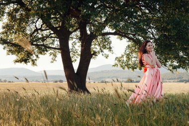 Yeşil yaz arka planında güzel aktif özgür kız. Bir tarlada dans eden güzel pembe bir elbiseyle güçlü bir meşe ağacının yanında