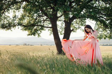 Yeşil yaz arka planında güzel aktif özgür kız. Bir tarlada dans eden güzel pembe bir elbiseyle güçlü bir meşe ağacının yanında