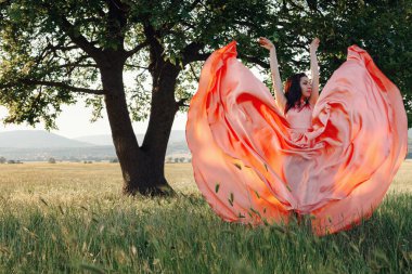 Yeşil yaz arka planında güzel aktif özgür kız. Bir tarlada dans eden güzel pembe bir elbiseyle güçlü bir meşe ağacının yanında