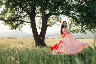 Yeşil yaz arka planında güzel aktif özgür kız. Bir tarlada dans eden güzel pembe bir elbiseyle güçlü bir meşe ağacının yanında