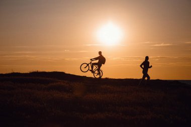 Bisikletli adamların silueti ve gün batımına doğru koşan bir kadın. Sağlıklı yaşam tarzı ve karantina sonlandırma fikri