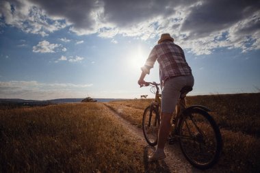 Yaz tatilinde bisikletle gezen genç çekici bir adam gün batımında toprak yolda kırsalda, sağlıklı aktif hayat tarzı gezgini, gömlek ve şapka giymiş.