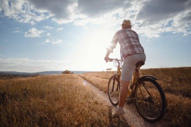 Yaz tatilinde bisikletle gezen genç çekici bir adam gün batımında toprak yolda kırsalda, sağlıklı aktif hayat tarzı gezgini, gömlek ve şapka giymiş.