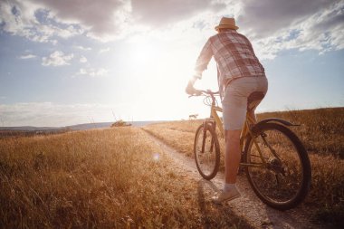 Yaz tatilinde bisikletle gezen genç çekici bir adam gün batımında toprak yolda kırsalda, sağlıklı aktif hayat tarzı gezgini, gömlek ve şapka giymiş.