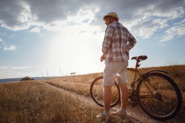 Yaz tatilinde bisikletle gezen genç çekici bir adam gün batımında toprak yolda kırsalda, sağlıklı aktif hayat tarzı gezgini, gömlek ve şapka giymiş.