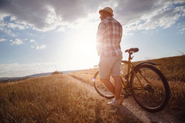 Yaz tatilinde bisikletle gezen genç çekici bir adam gün batımında toprak yolda kırsalda, sağlıklı aktif hayat tarzı gezgini, gömlek ve şapka giymiş.