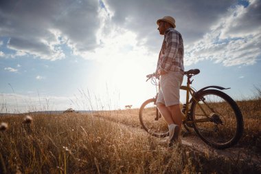 Yaz tatilinde bisikletle gezen genç çekici bir adam gün batımında toprak yolda kırsalda, sağlıklı aktif hayat tarzı gezgini, gömlek ve şapka giymiş.