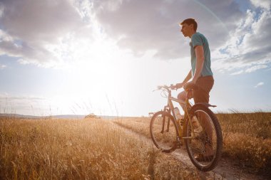 Günbatımında bisikletle gezen yakışıklı bir adam, sağlıklı, aktif bir hayat tarzı gezgini. Kırsalda gün batımında toprak yolda
