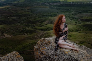 Kızıl kadın uçurumun kenarında oturuyor ve yaklaşan fırtınaya bakıyor, son ışığa bakıyor. Rüzgarda uçuşan saçlar. Dağ manzarasına hayran olan bir kız. Kırım, Klimentieva Dağı