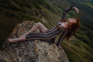 Kızıl kadın uçurumun kenarında oturuyor ve yaklaşan fırtınaya bakıyor, son ışığa bakıyor. Rüzgarda uçuşan saçlar. Dağ manzarasına hayran olan bir kız. Kırım, Klimentieva Dağı